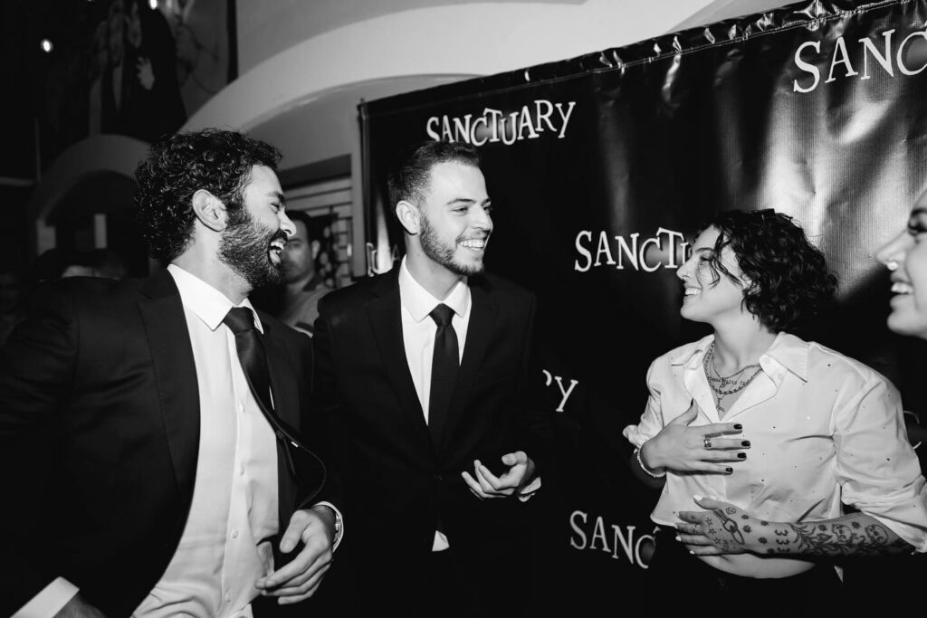 Three people in suits laughing and chatting at a black-tie event with a Sanctuary backdrop behind them.
