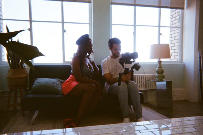 A man records video with a camera on a stabilizer while a woman sits on a sofa beside him in a bright living room.