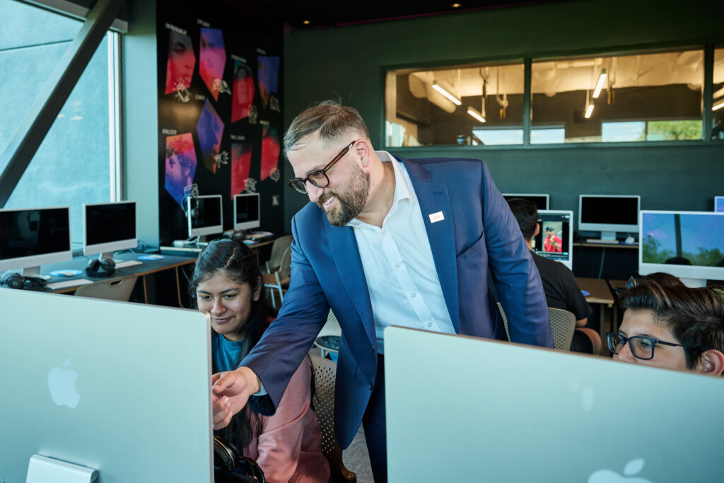 Man in a blue suit helps students at desktop computers in a modern computer lab classroom.
