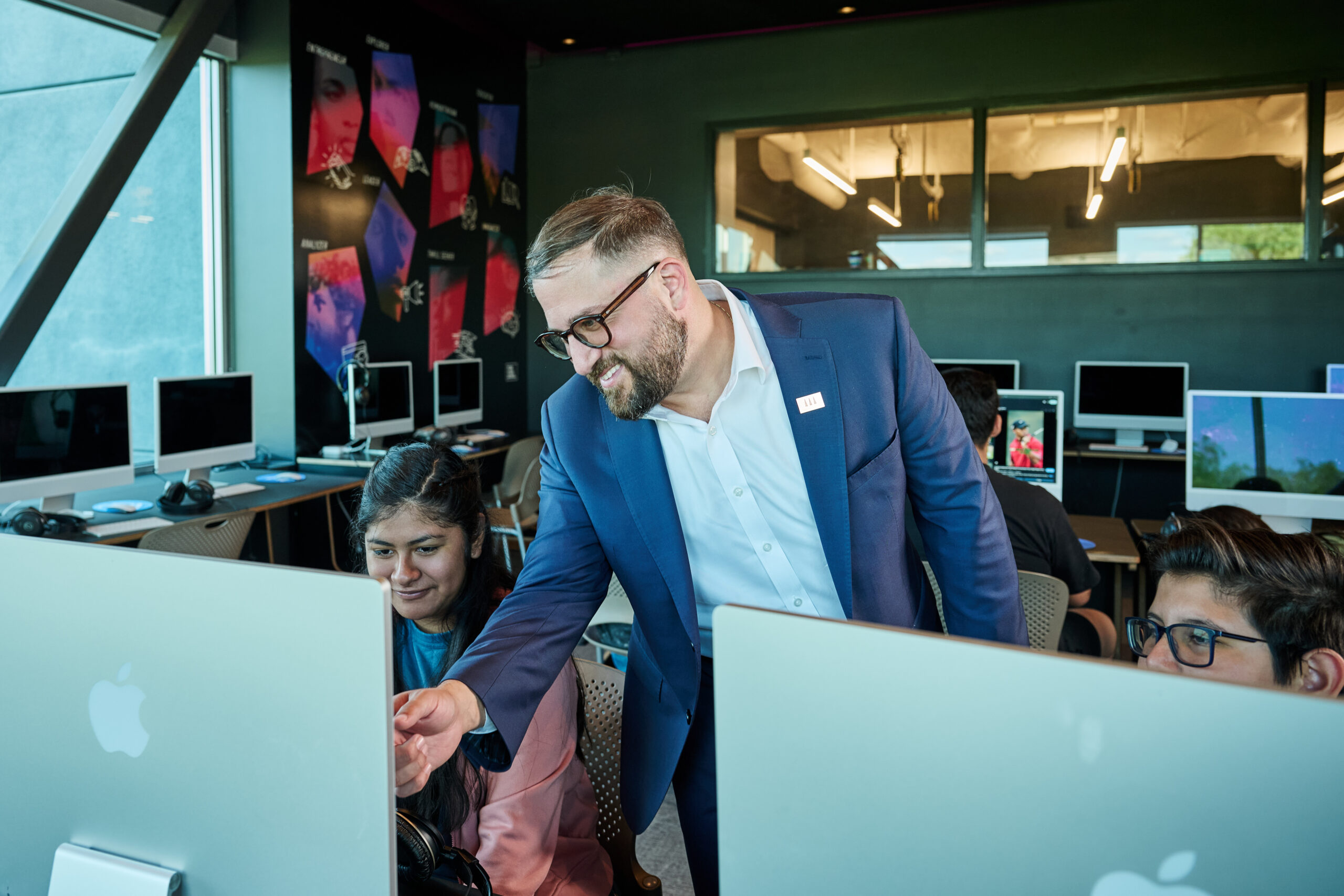 Man in a blue suit helps students at desktop computers in a modern computer lab classroom.