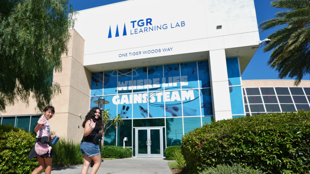 Two young women walk toward the glass-front entrance of TGR Learning Lab, waving at the camera with greenery and blue sky around them.