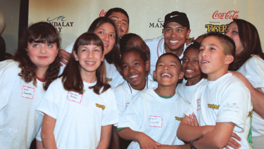 Group of smiling kids and teens in white T-shirts posing together at a sponsored charity event backdrop.