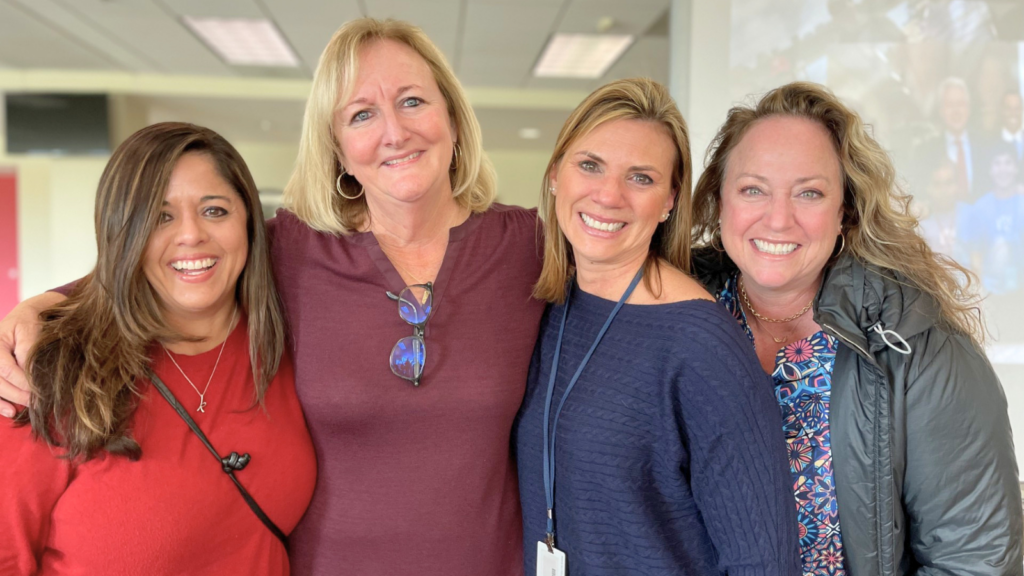 Four women stand close together indoors, smiling at the camera, arms around each other in a friendly group portrait.