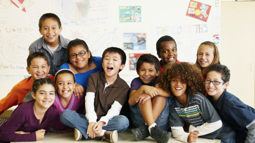 Group of diverse children smiling and posing together in a classroom with colorful posters on the wall behind them.