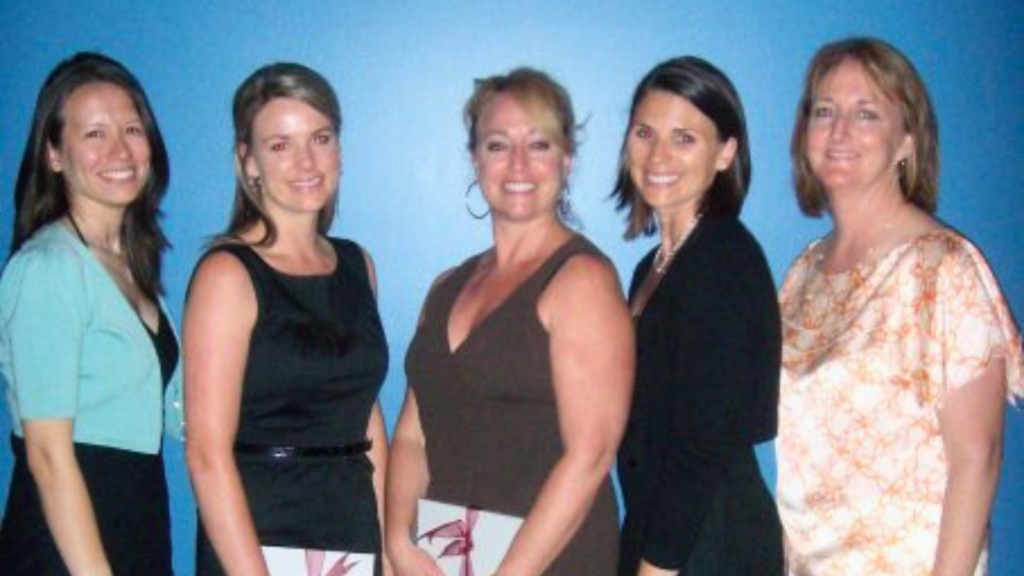 Five women stand in a row against a blue wall, smiling at the camera (group photo).