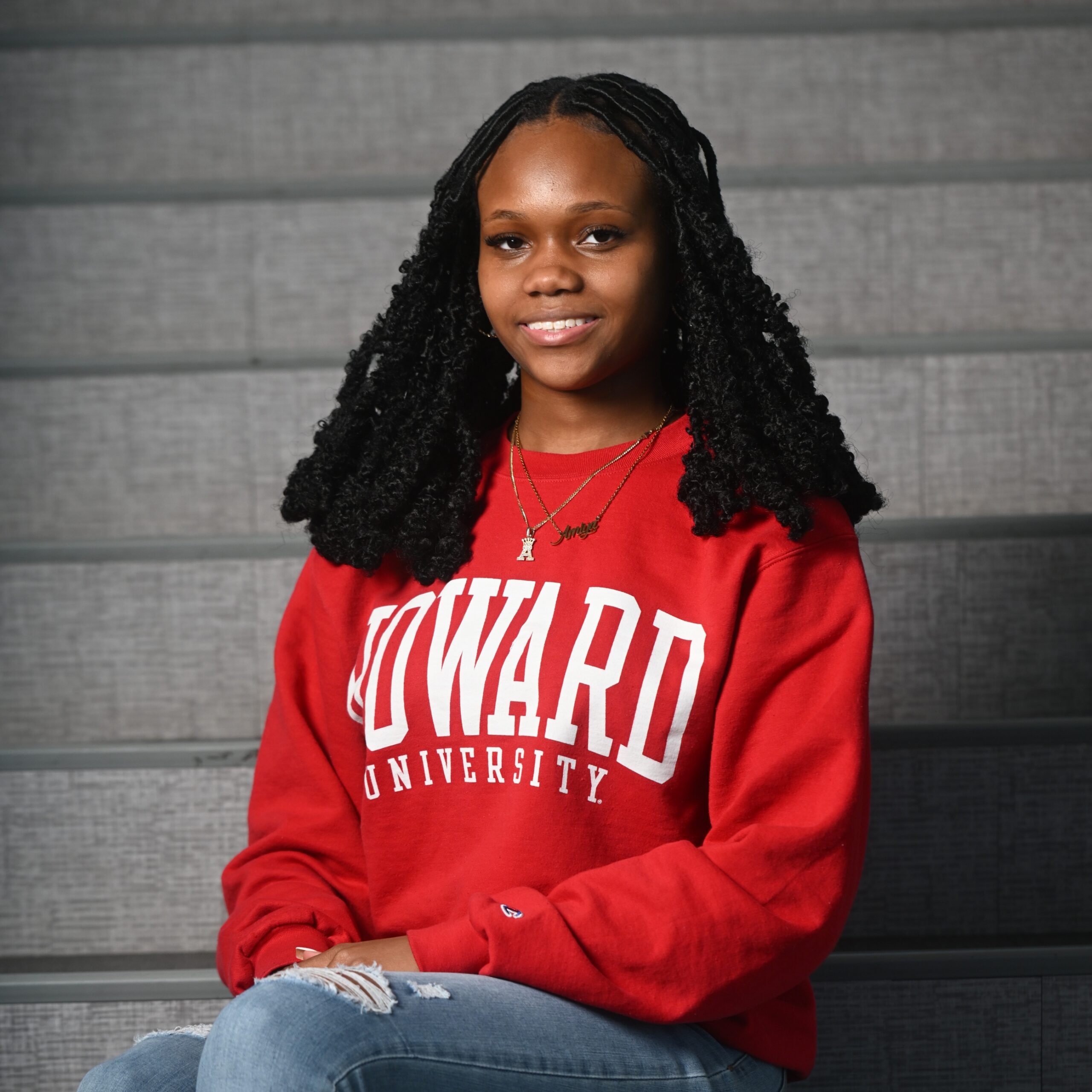 Portrait of a person sitting on gray bleachers, wearing a red sweatshirt with white 'UNIVERSITY' lettering and layered necklaces.