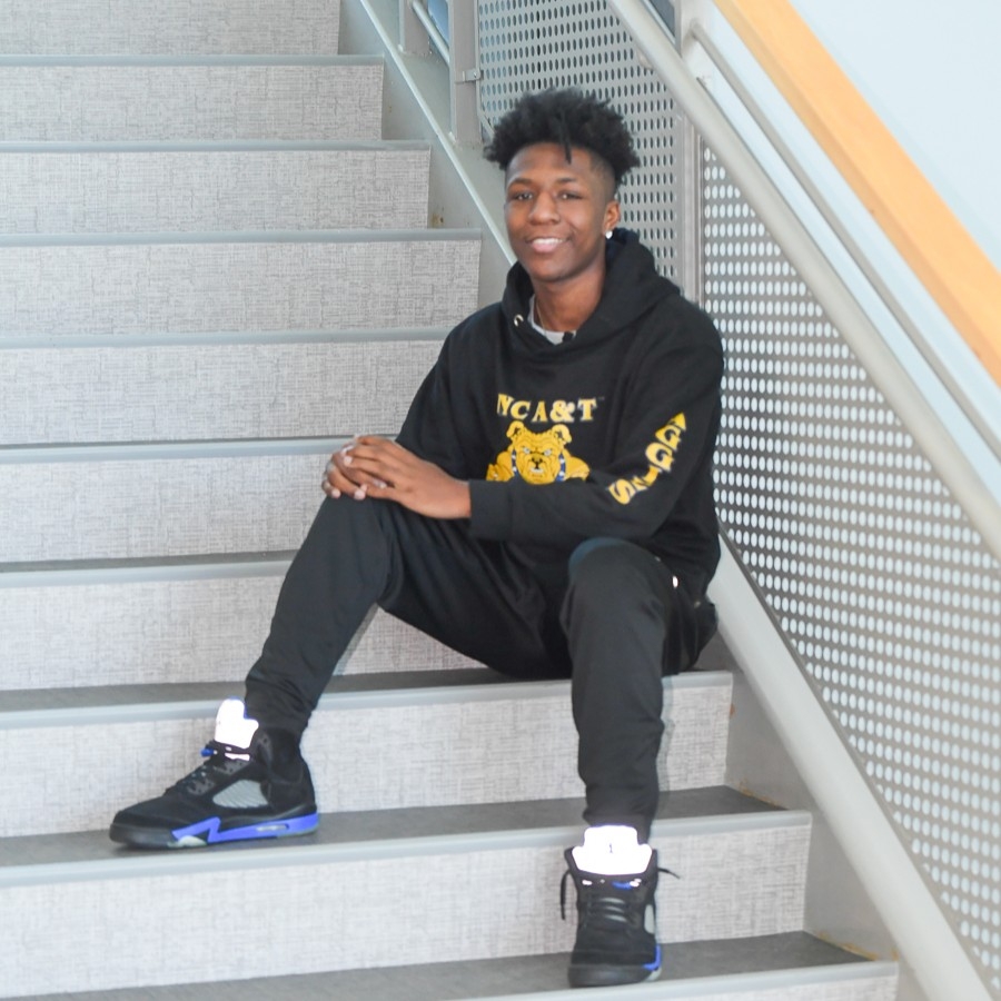 Young man sitting on indoor stairs, wearing a black hoodie with a yellow tiger logo and black sneakers.