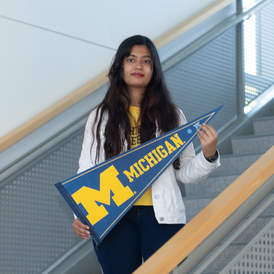 Woman standing on an indoor staircase holding a blue Michigan pennant with a large yellow M on it.