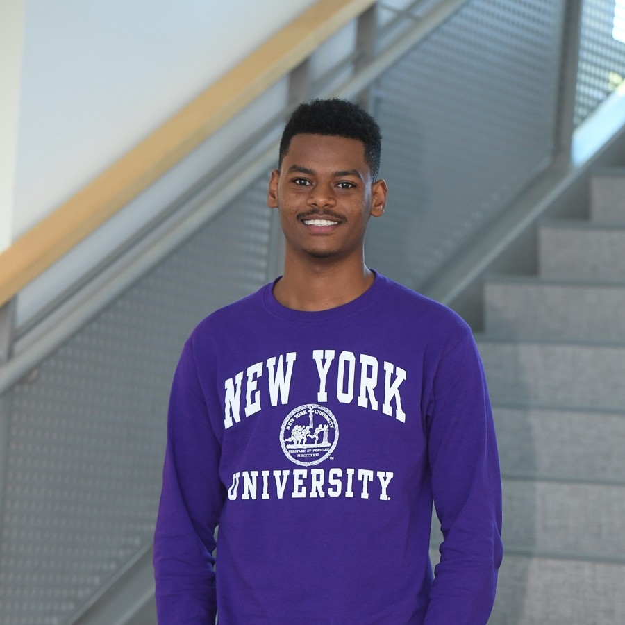 Portrait of a young man in a purple New York University sweatshirt standing on a stairwell with a metal railing behind him.