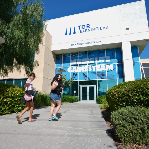 Two girls walk toward the entrance of TGR Learning Lab on a sunny day, with a glass façade and palm trees nearby; the building sign reads 'TGR Learning Lab' and 'One Tiger Woods Way.'