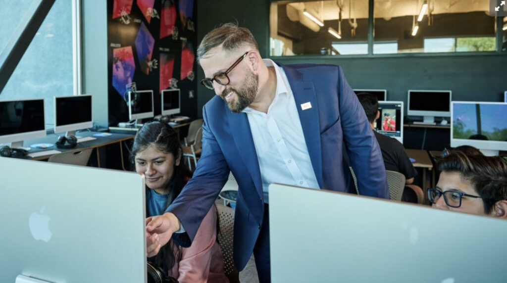 Man in a blue suit helps a student at an Apple iMac in a computer lab classroom