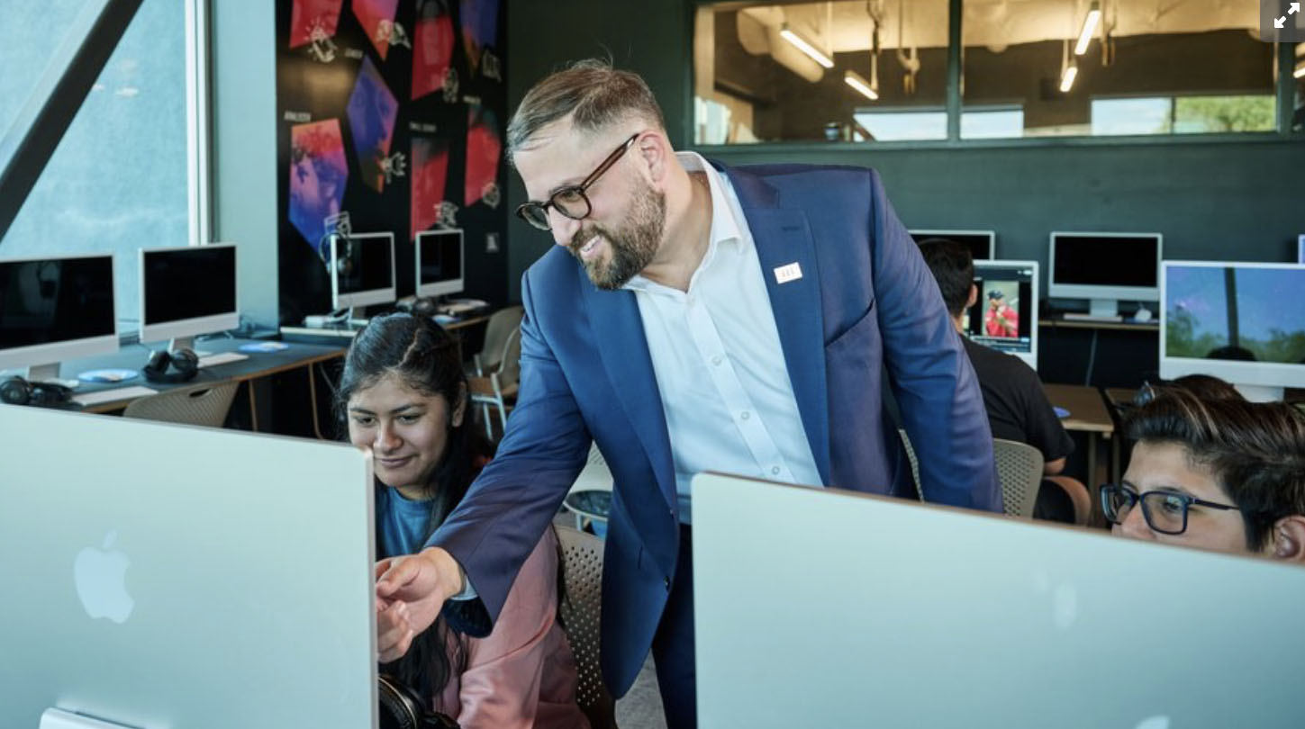 Man in a blue suit helps a student at an Apple iMac in a computer lab classroom