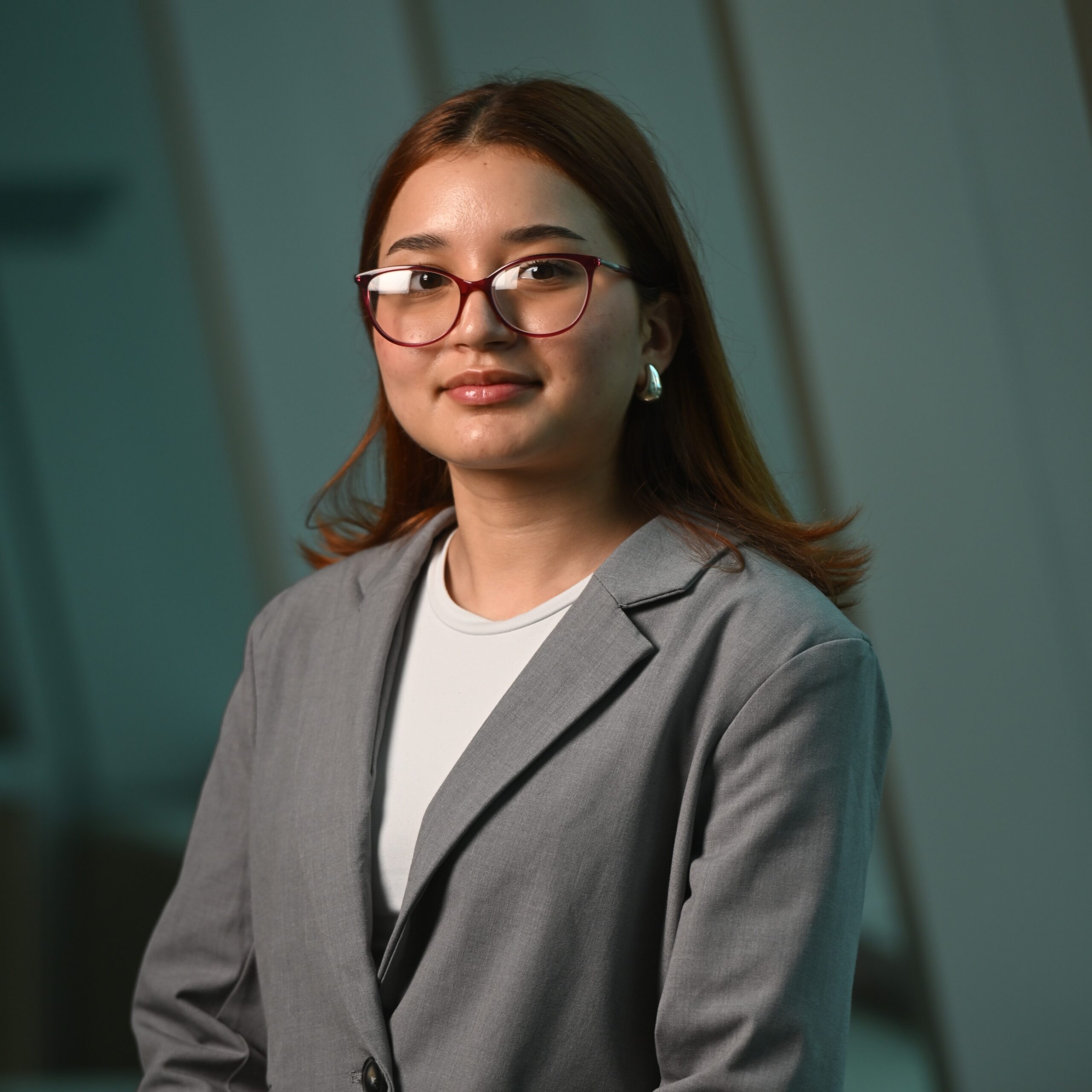 Woman wearing red-framed glasses and a gray blazer, looking slightly to the side indoors with a neutral expression.